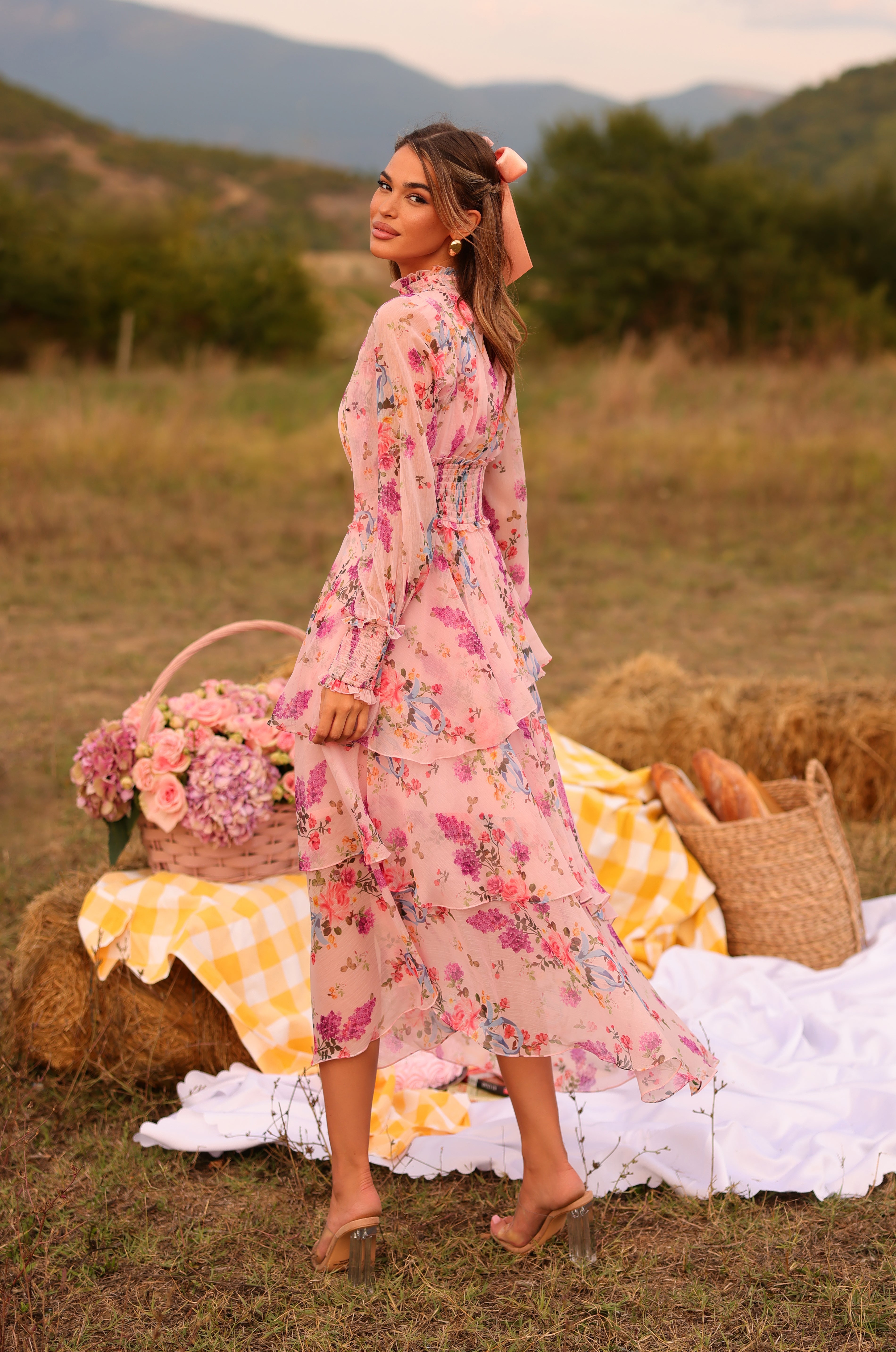Wearing the Bellaria Long Sleeve Blush Pink Floral Maxi Dress, a woman stands on grass by a picnic setup with hay bales, flowers, and bread, surrounded by trees and mountains.